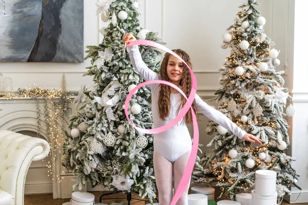 A beautiful little gymnast in a white sports dress doing rhythmic gymnastics exercises spirals with an artistic ribbon in a fitness class during the Christmas season. Sports, training, stretching