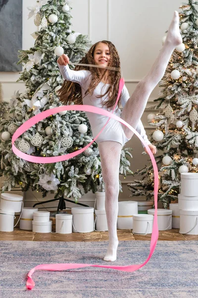 A beautiful little gymnast in a white sports dress doing rhythmic gymnastics exercises spirals with an artistic ribbon in a fitness class during the Christmas season. Sports, training, stretching