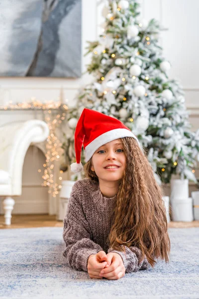 迷人的卷发小女孩头戴圣诞礼帽 微笑着看着明亮的圣诞装饰品工作室里的摄像机 — 图库照片