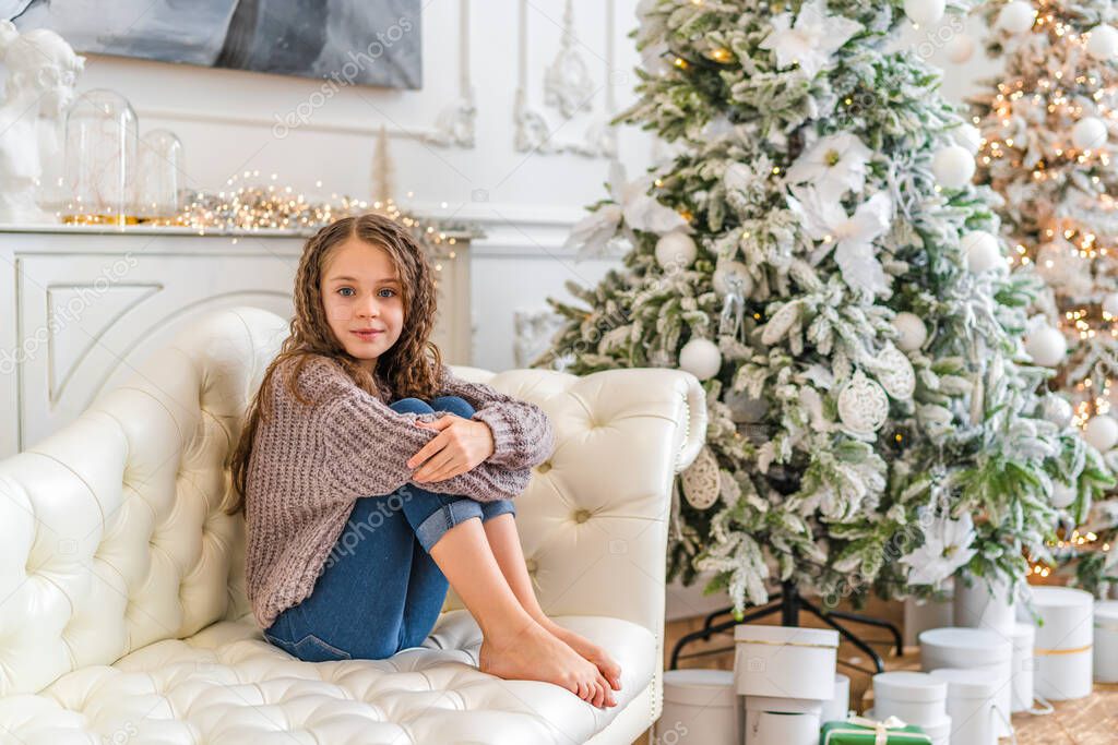 A little girl dressed in a warm knitted sweater sits on the sofa next to the Christmas tree