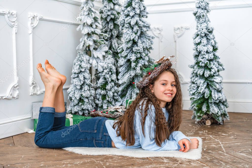 A little girl in a cozy blue sweater lies on the carpet under the Christmas tree