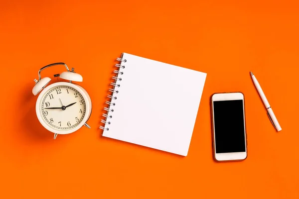 An empty blank notepad for writing on an orange background and an alarm clock. The concept of planning and getting things done. Minimalism