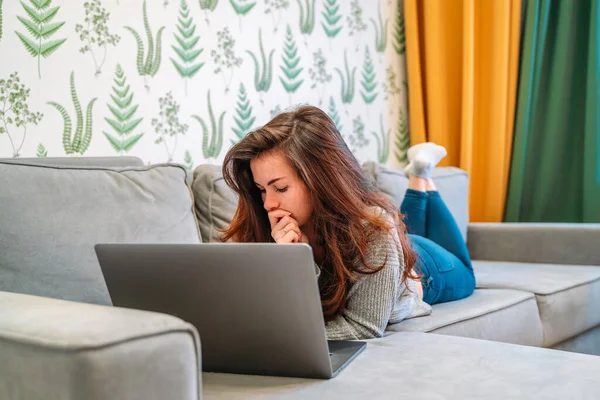 A modern young woman relaxes on the sofa in the living room using a laptop. Work from home and education.
