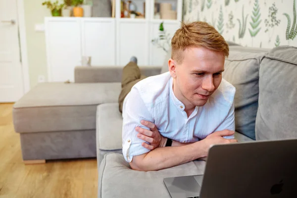 A modern young man relaxes on the sofa in the living room using a laptop. Work from home and education.