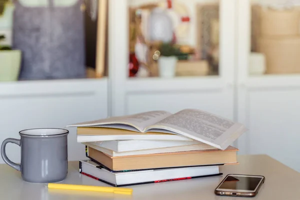 A stack of books, a mug of tea, and a mobile phone on a white table. A bookcase with books in the background in a modern interior. The concept of training and education.