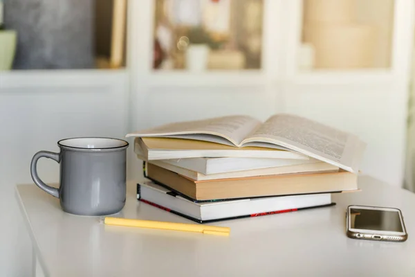A stack of books, a mug of tea, and a mobile phone on a white table. A bookcase with books in the background in a modern interior. The concept of training and education.
