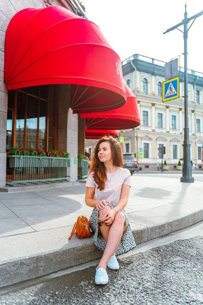 A young woman in a skirt sits on the sidewalk under the red awning of the famous Astoria Hotel in St. Petersburg, on a sunny day. Saint Petersburg, Russia - 05 Apr 2021
