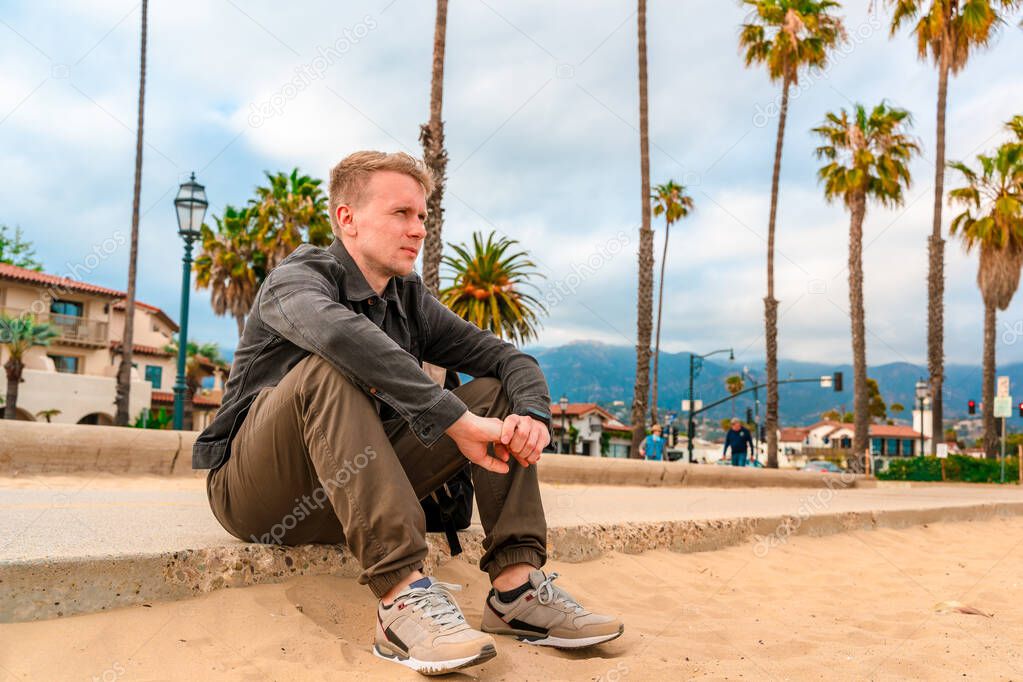 A man sits on a boardwalk along palm trees in Santa Barbara, California
