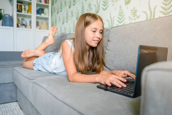 小さな女子高生が家で勉強していて ノートパソコンを持ってソファの上に横たわっている 遠隔オンライン教育 — ストック写真