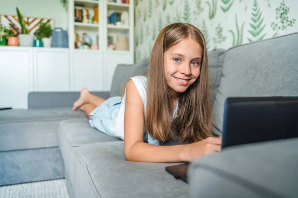 小さな女子高生が家で勉強していて ノートパソコンを持ってソファの上に横たわっている 遠隔オンライン教育 — ストック写真