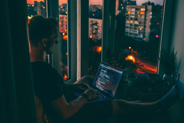 A young programmer is coding on a laptop in the dark sitting on a window with a view of the lights of the night city, colored lighting in the room, home decor