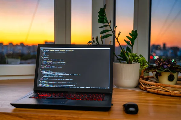 The code is on a laptop on a wooden table in front of the window  in the dark with a view of the lights of the night city, color lighting in the room, home decor