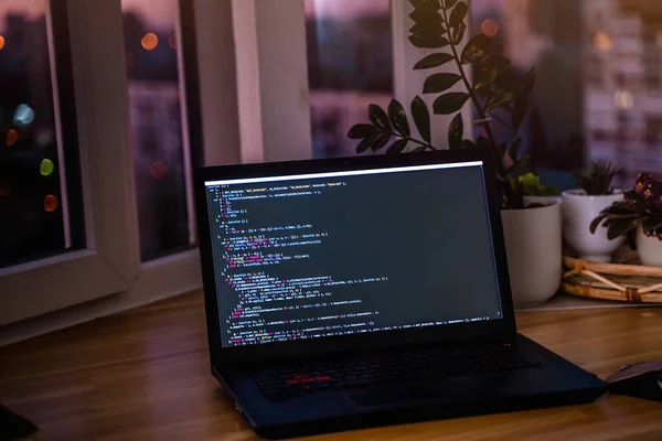 The code is on a laptop on a wooden table in front of the window  in the dark with a view of the lights of the night city, color lighting in the room, home decor