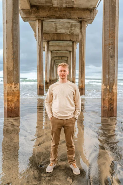 Young man is standing under pier in La Jolla Shores at the cloudy time. Symmetrical Pier in San Diego, California