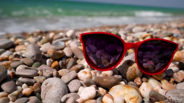Сонцезахисні Окуляри Вигляді Сердець Лежать Гальковому Пляжі Морськими Хвилями Задньому — стокове відео
