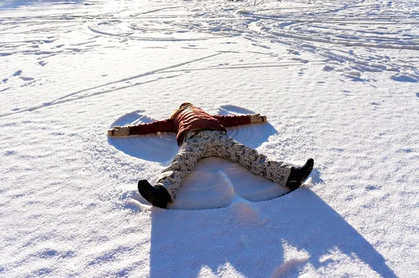 Young woman lying in the fresh snow and making a snow angel. Winter walking and fun concept, holidays and vacation