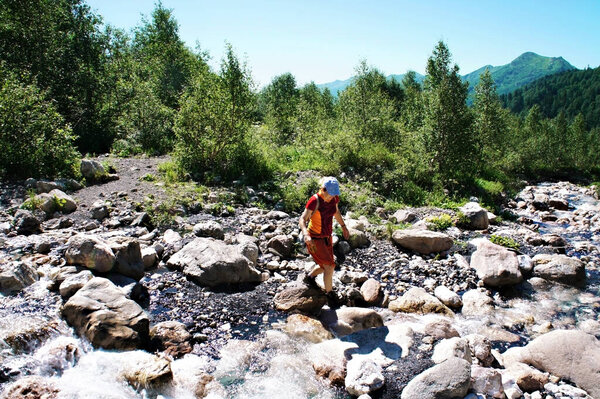 Young Caucasian woman tourist in red clothes hiking through mountain river on stones against background of green vegetation in Caucasus mountains in summer. Active lifestyle, travel destination