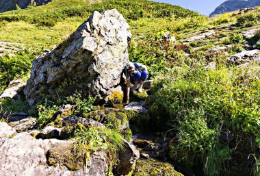 Şapkalı genç bayan yürüyüşçü, Kafkasya Dağları 'nın arka planındaki büyük kayanın yakınındaki dağ nehrinden gelen soğuk suya dokunur.