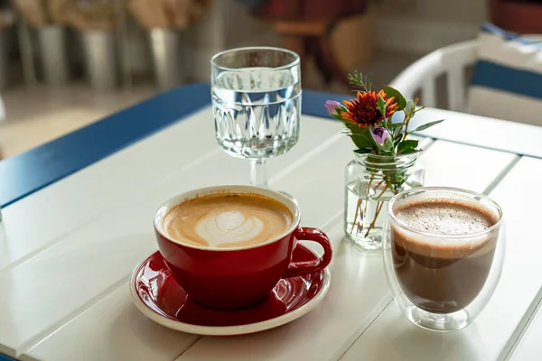 赤マグカップのカプチーノ ガラスカップのカカオ カフェのテーブルの上のガラス瓶の中の水と小さな花束の花 装飾品 ホットドリンク 静物画 コーヒータイム — ストック写真