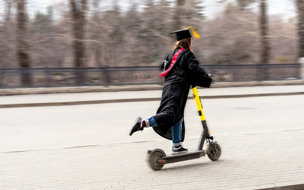 Üniversiteden mezun olduktan sonra siyah mezuniyet elbisesi ve sarı püsküllü siyah şapkalı, elektrikli scooter süren mutlu bir genç kadın. — Stok fotoğraf