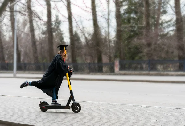 大学毕业后，身穿黑色毕业礼服、头戴黑色毕业帽、头戴黄色花冠、骑电动车的快乐年轻女性走在大街上 图库图片