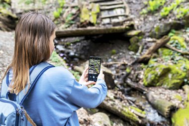 Young blond woman in blue sweatshirt taking photo of beautiful mountain river on asmartphone in mossy forest, hiking, digital detox, beauty in nature, active lifestyle