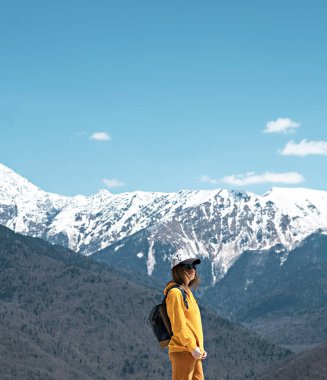 side view Young blond woman in yellow hoodie, cap, with backpack on background of snow capped Caucasus mountains. Hiking and travel, active lifestyle, copy space
