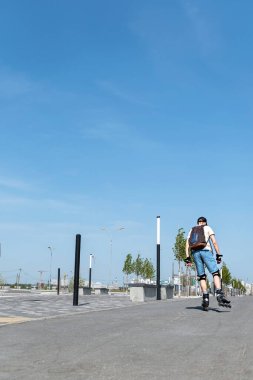 Mavi şortlu, sırt çantalı, koruyucu ekipmanlı, yaz aylarında asfalt yolda paten süren, patenli, açık hava aktiviteli, sağlıklı aktif yaşam tarzı olan genç bir adam.