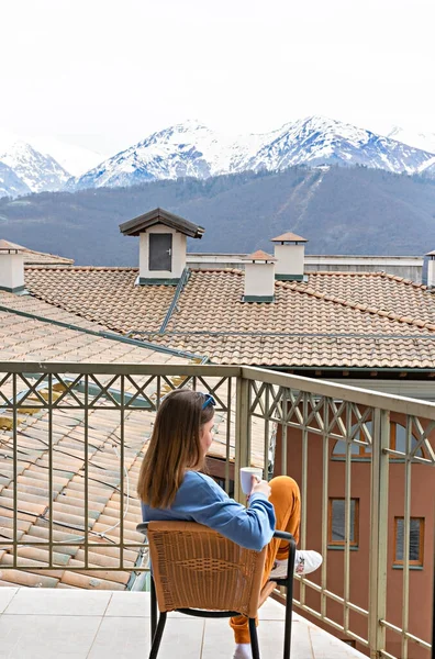 Rear view of blond young woman in blue sweatshirt sitting with mug of drink in chair on terrace and looking at mountain view with snowy peaks, Relaxation and enjoying the moment, selective focus