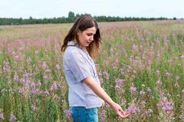 Mor gömlekli, gülümseyen güzel sarışın kadın çayırlarda yürüyor ateş otları ve dokunaklı çiçeklerin arasında, doğadaki güzellik seçici odak noktası.