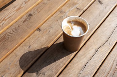 Latte or cappuccino with plant milk in beige paper cup on wooden striped table outside, drinking coffee, light and shadow .