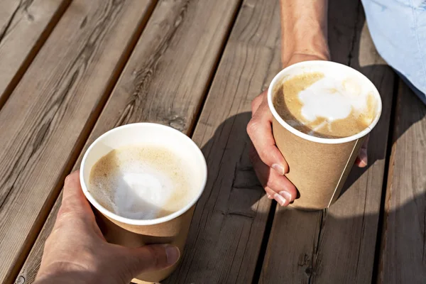 Hands of two people holding beige paper cups of cappuccino on a wooden striped table outside, drinking coffee, meeting friends