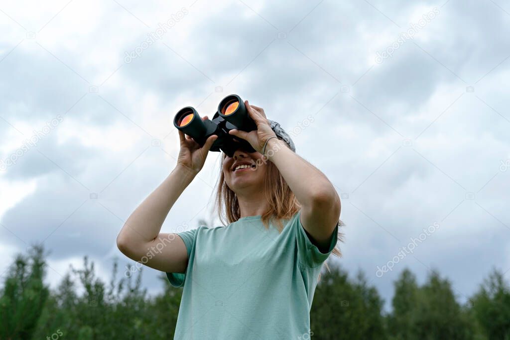 smiling Young blonde woman bird watcher in cap and blue t-shirt looking through binoculars at cloudy sky in summer forest ornithological research Birdwatching, zoology, nature, observation of animals 