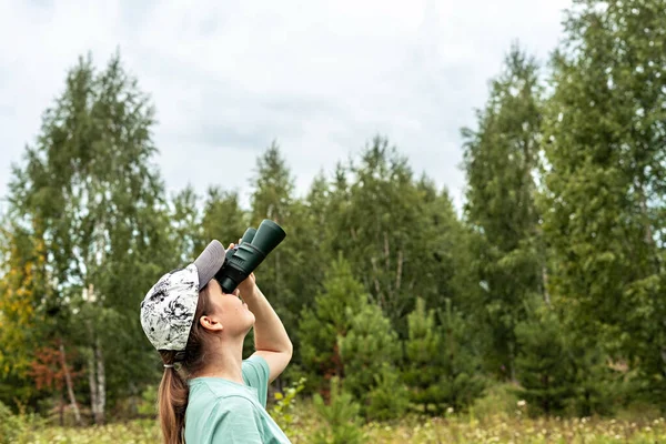 Young blonde woman bird watcher in cap and blue looking through binoculars at cloudy sky in summer forest ornithological research Birdwatching, zoology, nature, observation of animals Ornithology