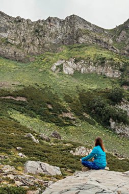 Meditasyon yapan mavi elbiseli genç bir kadının yeşil dağ vadisinde taşın üzerinde oturuşu, doğada rahatlama, bilinç ve farkındalık.