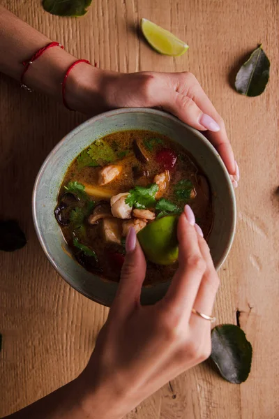 female hands squeezing lime to soup with shrimps, vegetables, and pieces of fish on the table