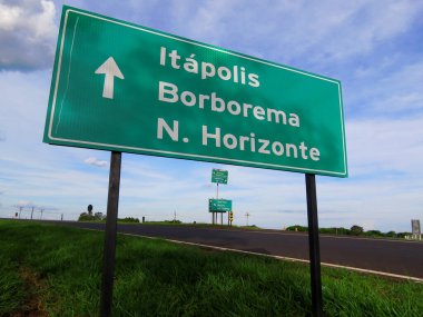 Metal plaka, Brezilya 'da canlı renklerde bir kırsal yolun kenarına yerleştirilmiş büyük beyaz bir ok ile üç şehrin yönünü verir (Portekizce gösterilir).