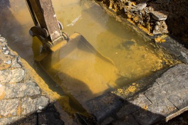 Paslı kazıcı, sokaktaki kanalizasyon sisteminin kirli sularının altını kazıyor. Sahneyi oluşturan parlak bir ışık ve gölgeler var.