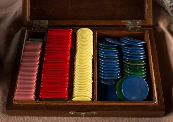 Multiples plastic poker chips (or used for another game) inside beautiful old crafted wooden box. Different colors of chips stacked all together