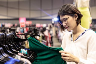 Asyalı genç, kısa saçlı, gözlüklü güzel bir kadın alışveriş merkezinde satın almak için askıdaki gömlek koleksiyonuna bakıyor.