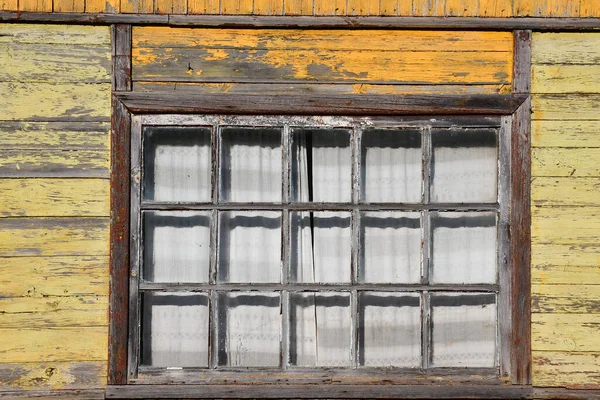 A fragment of a wall made of planks with peeling yellow paint. A fifteen-pane window with an old wooden frame.