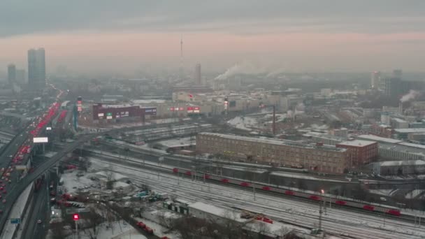 Příměstský vlak projíždí pod mostem, na kterém se v zimě zasekává dopravní zácpa na pozadí městské krajiny v mlze. letecký pohled — Stock video