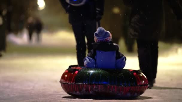 Ένα μικρό παιδί γονείς βόλτα ένα χειμώνα φουσκωτό έλκηθρο κατά τη διάρκεια μιας χιονόπτωση στο πάρκο νύχτα — Αρχείο Βίντεο