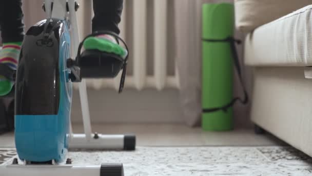 Beine, die einen Heimtrainer zu Hause in einem Raum in die Pedale treten, Nahsicht. Dolly-Kamerafahrt — Stockvideo
