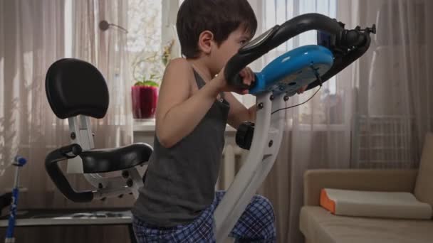 Little boy in home clothes pedals an exercise bike at home in the room — Stock Video