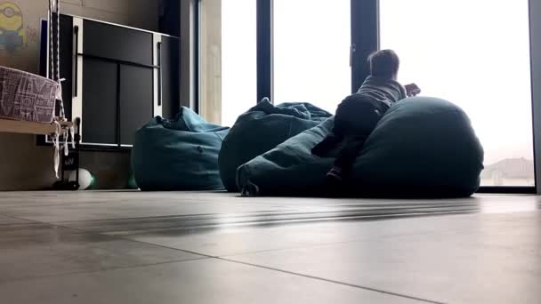 Een robot stofzuiger reinigt de kamer terwijl een jongetje op een poef ligt bij het panoramische raam — Stockvideo