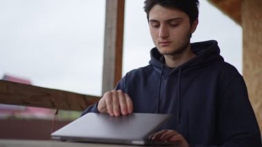 Genç bir adam bir terasta otururken dizüstü bilgisayarında resim gibi kırsal bir arazinin arka planına karşı yazı yazıyor. Adam köyde bir dizüstü bilgisayarda çalışıyor..