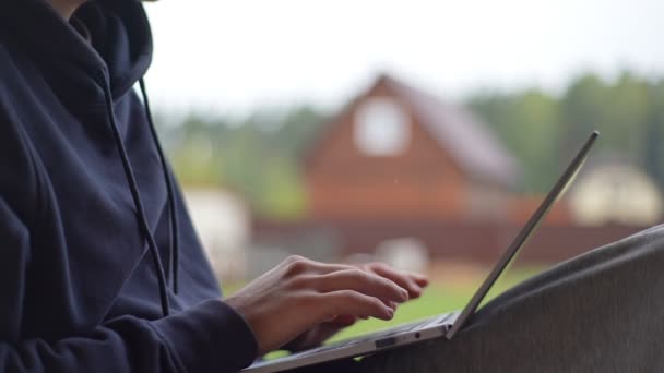 Ένας νεαρός πληκτρολογεί σε ένα φορητό υπολογιστή, ενώ κάθεται σε μια βεράντα με φόντο ένα γραφικό αγροτικό τοπίο. Ο τύπος δουλεύει σε ένα λάπτοπ στο χωριό.. — Αρχείο Βίντεο