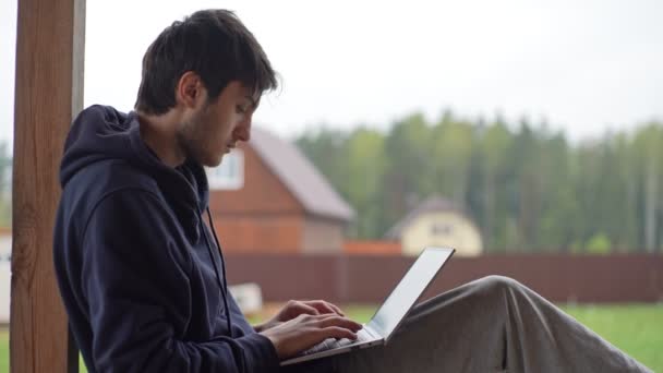Un joven está escribiendo en un ordenador portátil mientras está sentado en una terraza con el telón de fondo de un pintoresco paisaje rural. El tipo trabaja en un portátil en el pueblo.. — Vídeo de stock