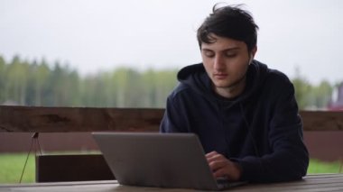 Genç bir adam bir terasta otururken dizüstü bilgisayarında resim gibi kırsal bir arazinin arka planına karşı yazı yazıyor. Adam köyde bir dizüstü bilgisayarda çalışıyor..